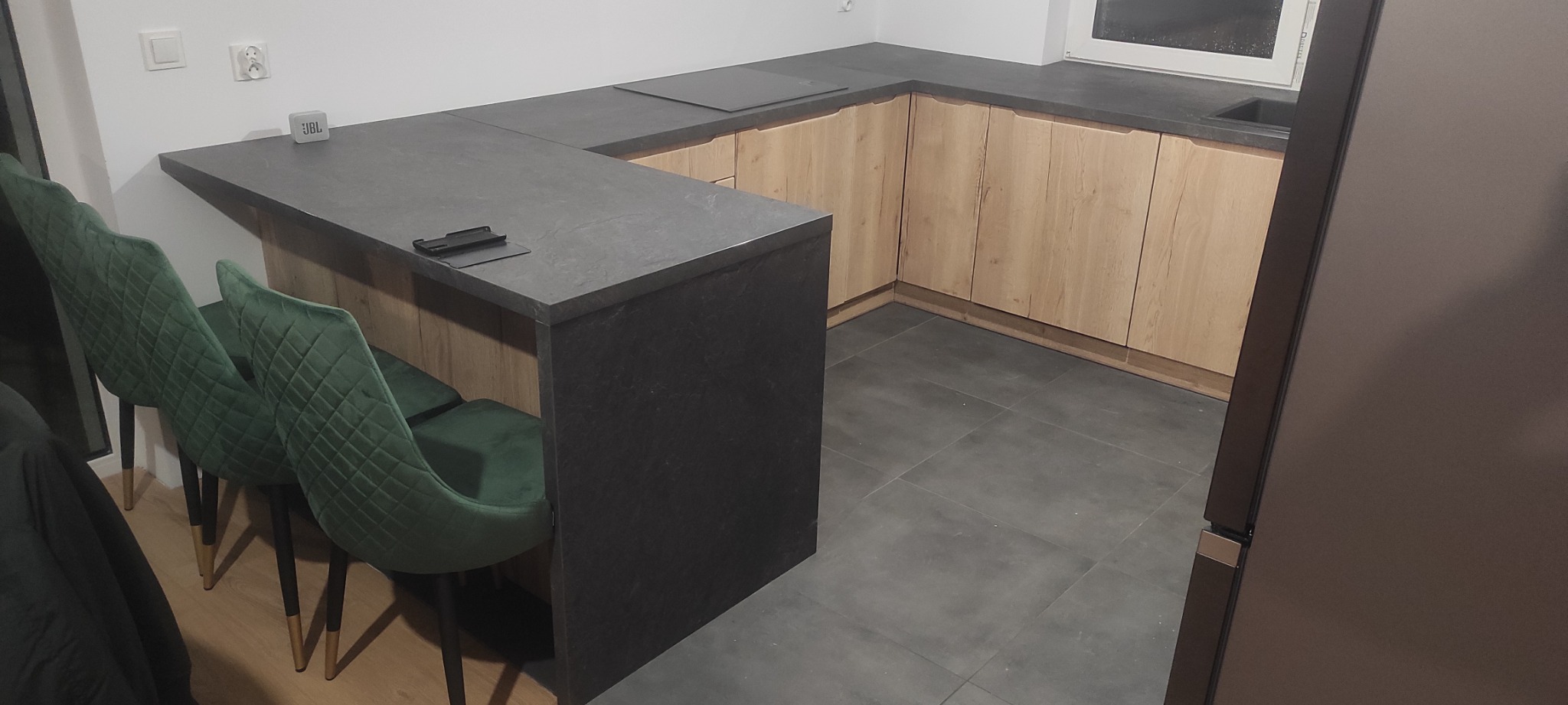 Nowoczesna kuchnia z drewnianymi szafkami, ciemnoszarym blatem i barkiem, uzupełniona o dwie butelkowe, pikowane krzesła na złotych nóżkach, z widocznym głośnikiem JBL na blacie.