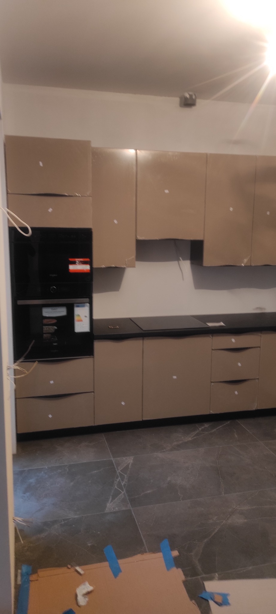 Kuchnia na etapie montażu z frontami zabezpieczonymi folią ochronną, widoczne urządzenia AGD i fragment podłogi.