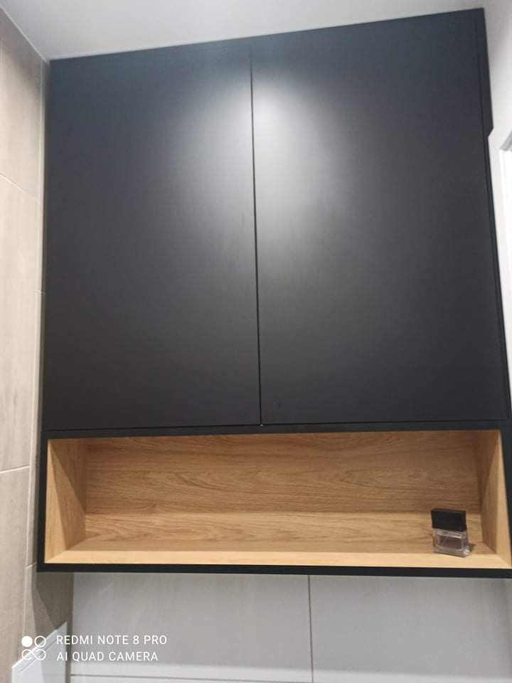 Ciemna szafka wisząca z drewnianą wnęką, na której stoi flakon perfum, na tle białych płytek.