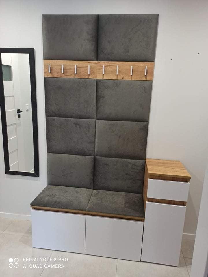 Panel tapicerowany z siedziskiem i szafkami w przedpokoju, z drewnianymi wstawkami i wieszakami, obok lustro w czarnej ramie.