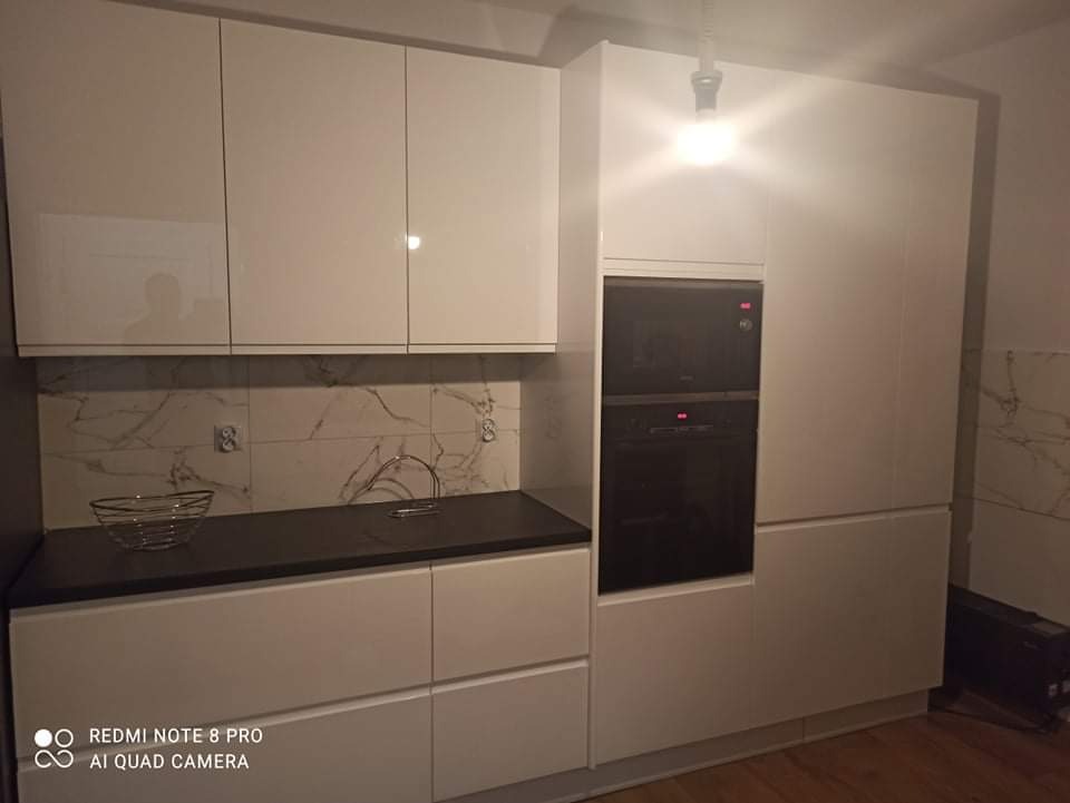 Nowoczesna kuchnia z białymi, lakierowanymi frontami, ciemnym blatem i marmurowym backsplash'em, wyposażona w piekarnik w zabudowie, oświetlenie halogenowe.