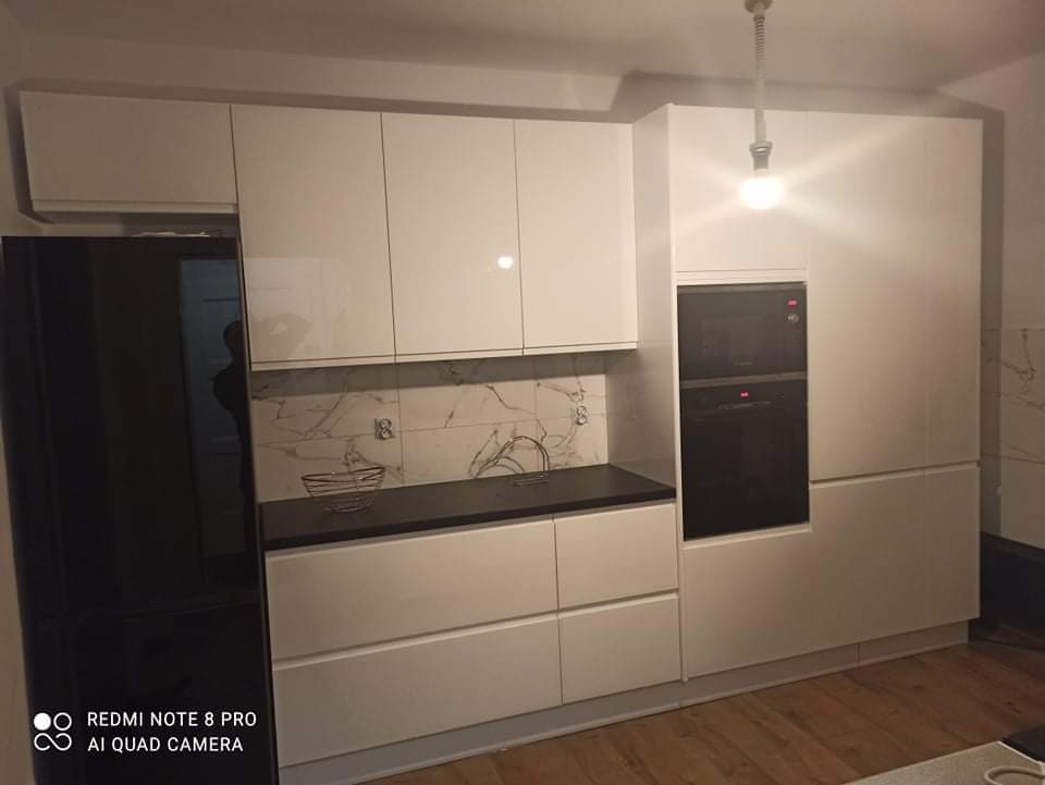 Nowoczesna, biała kuchnia z czarnym blatem i sprzętem AGD w zabudowie, widok z niskiego kąta, drewniana podłoga, marmurowe wykończenie ściany nad blatem.