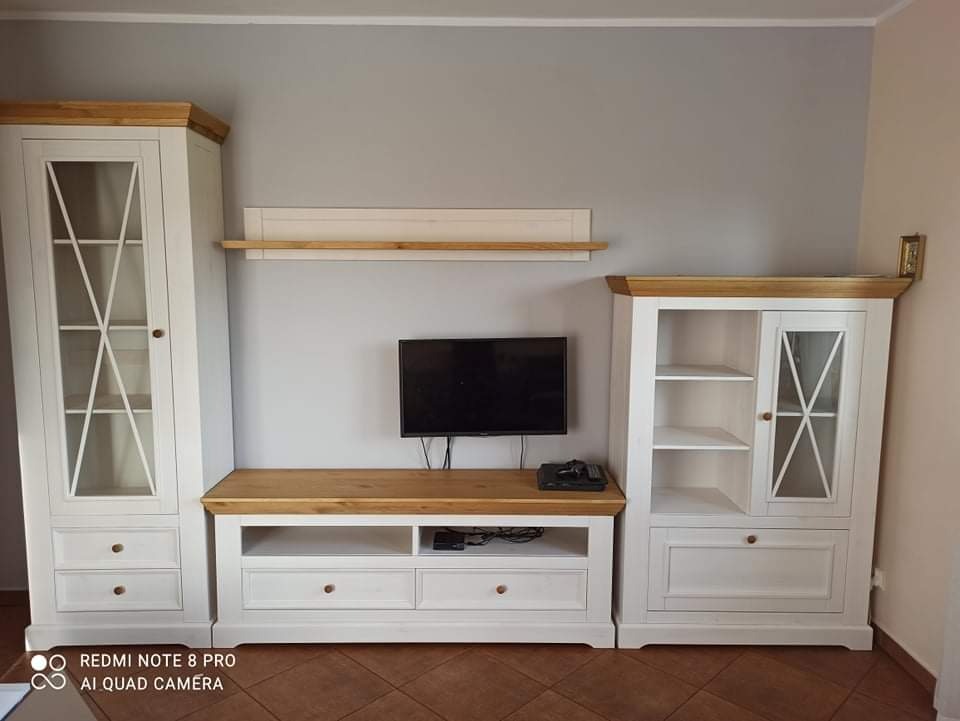 Zestaw mebli do salonu w stylu rustykalnym, składający się z dwóch białych witryn z drewnianymi blatami i przeszklonymi drzwiczkami, połączonych szafką RTV z szufladami i półkami, na ścianie...