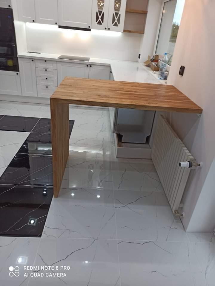 Nowoczesna kuchnia z białymi szafkami i blatem z jasnego drewna, tworzącym półwysep, z widocznym grzejnikiem pod blatem i błyszczącą podłogą w czarno-białą szachownicę.