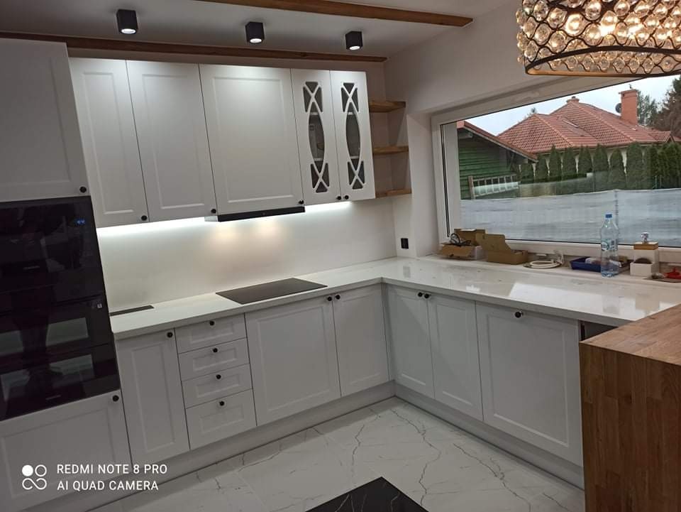 Jasna kuchnia z białymi szafkami, czarnym piekarnikiem i marmurową podłogą, widok na zewnątrz przez duże okno, ozdobna lampa wisząca.