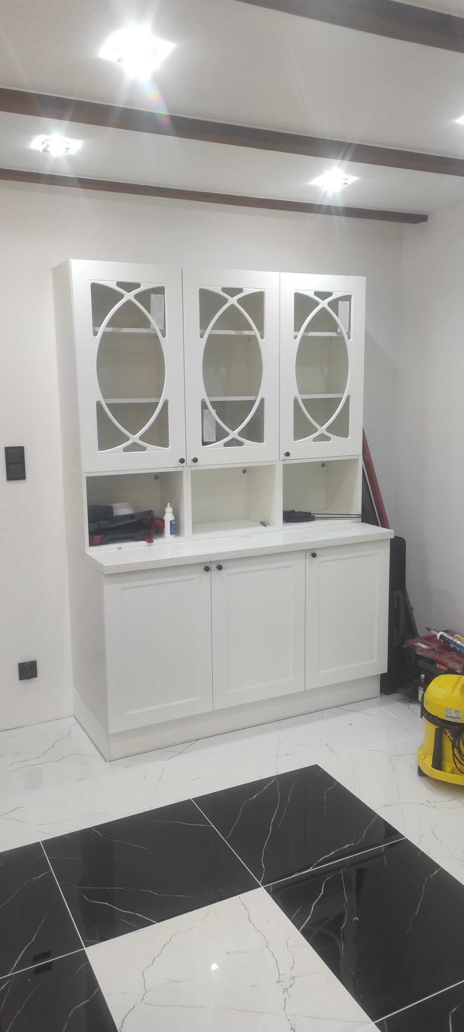 Biała szafka kuchenna z przeszklonymi drzwiczkami i dekoracyjnym wzorem, stojąca na czarno-białej marmurowej podłodze, z widocznymi narzędziami i akcesoriami na blacie.