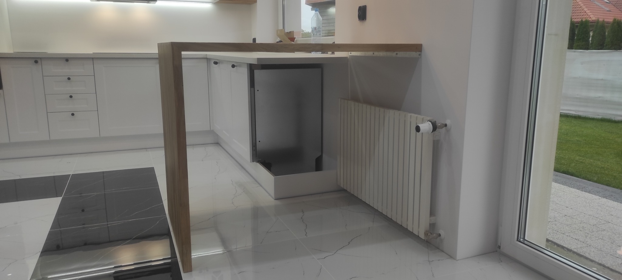 Nowoczesna, jasna kuchnia z białymi szafkami i blatem z jasnego drewna, wykończona marmurowymi płytkami podłogowymi w odcieniach bieli i szarości, z widocznym fragmentem grzejnika.