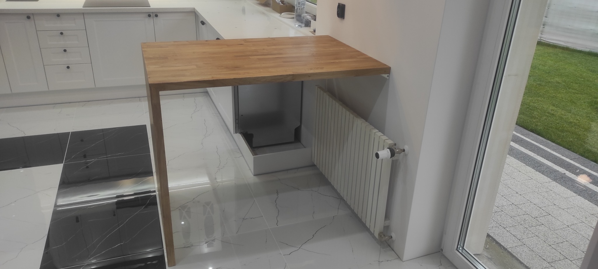 Kuchenny półwysep z blatem z jasnego drewna, wsparty na jednej nodze i przymocowany do ściany, z widocznym grzejnikiem i fragmentem białej zabudowy kuchennej na tle marmurowej podłogi i okna...