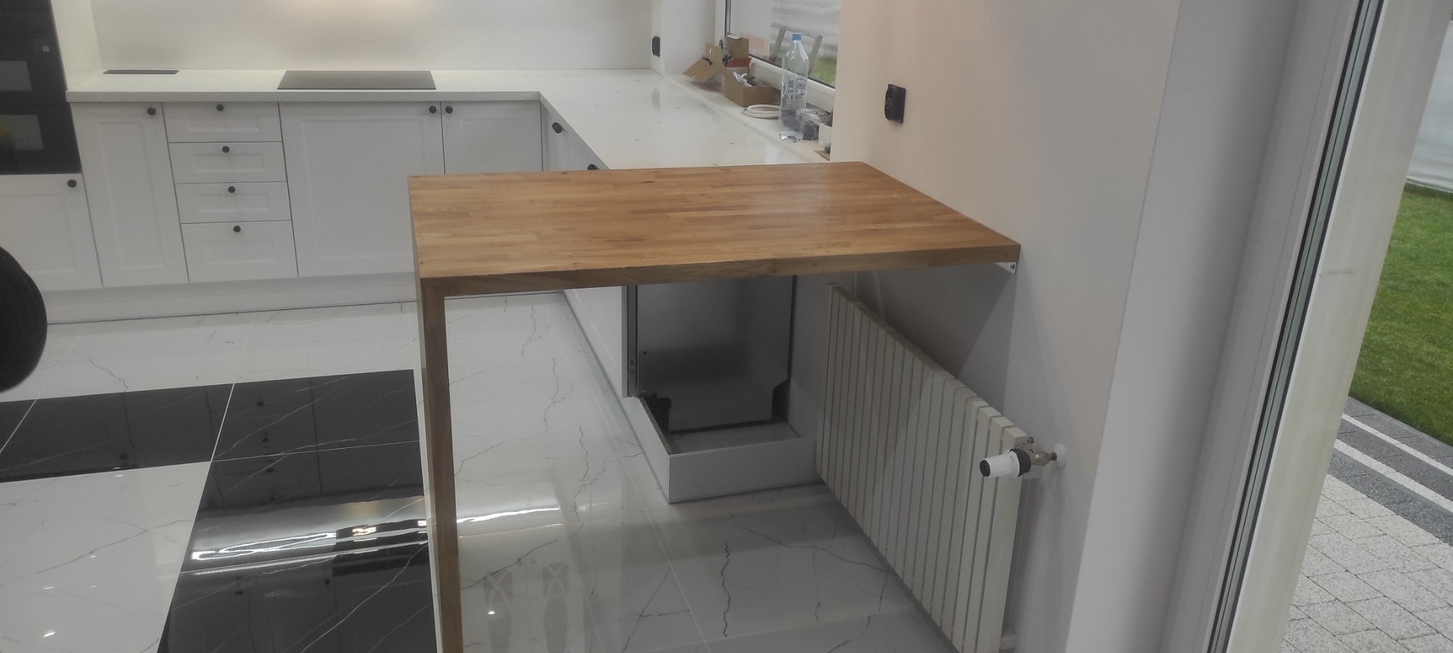 Drewniany blat przymocowany do ściany w nowoczesnej kuchni z białymi szafkami i czarno-białą podłogą, widok na ogród przez okno.