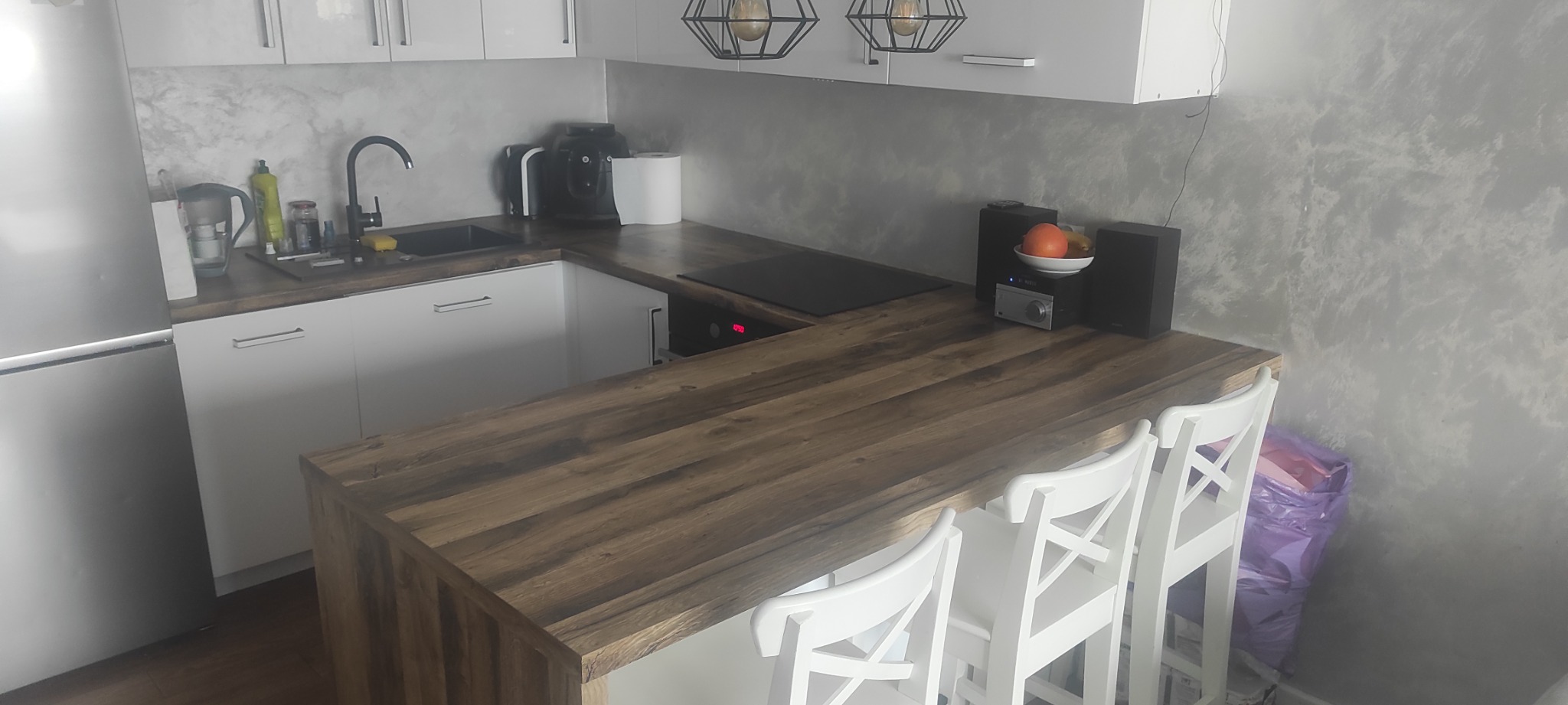 Nowoczesna kuchnia z białymi szafkami i drewnianym blatem z barkiem, uzupełniona o białe hokery, czarny zlew i baterię, oraz dekoracyjne lampy wiszące w stylu industrialnym.