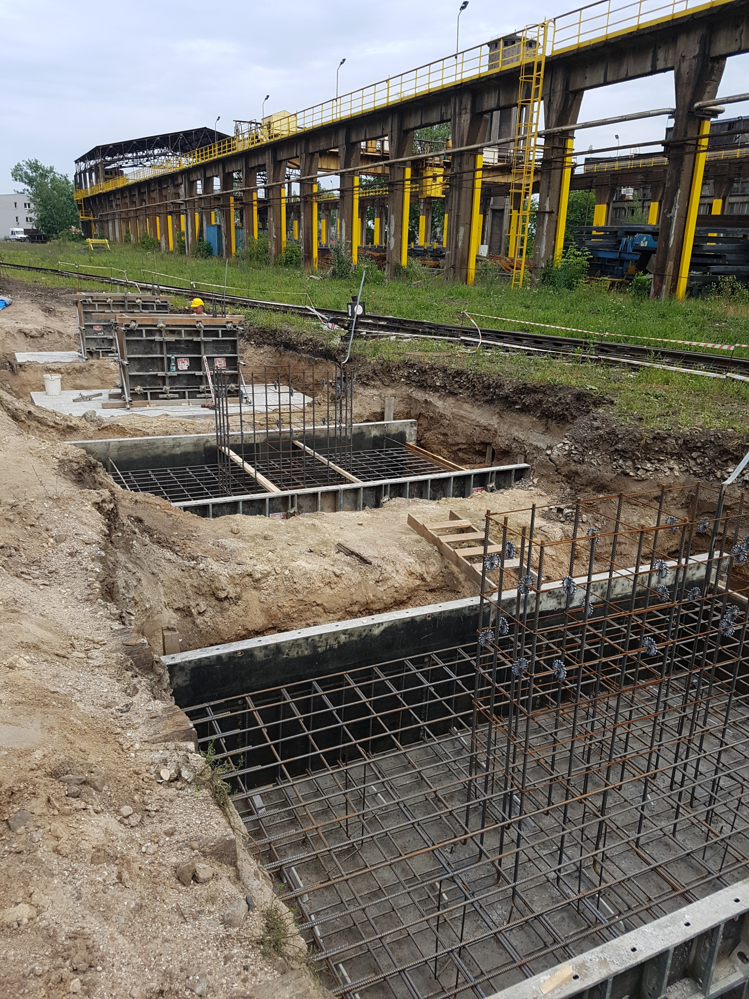 Przygotowanie zbrojenia i szalunków pod fundamenty w Konstancin-Jeziorna, widoczne metalowe konstrukcje i wykop pod budowę, w tle stara hala przemysłowa.