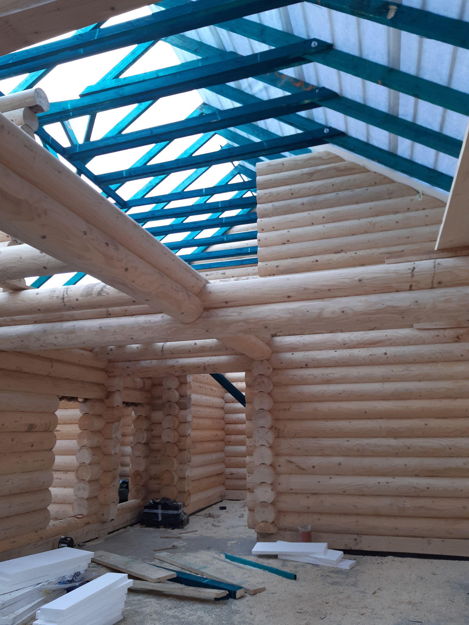 Wnętrze surowego domu z bali w trakcie budowy, widoczne drewniane ściany z okrąglaków, niebieska konstrukcja dachu i materiały izolacyjne na podłodze.