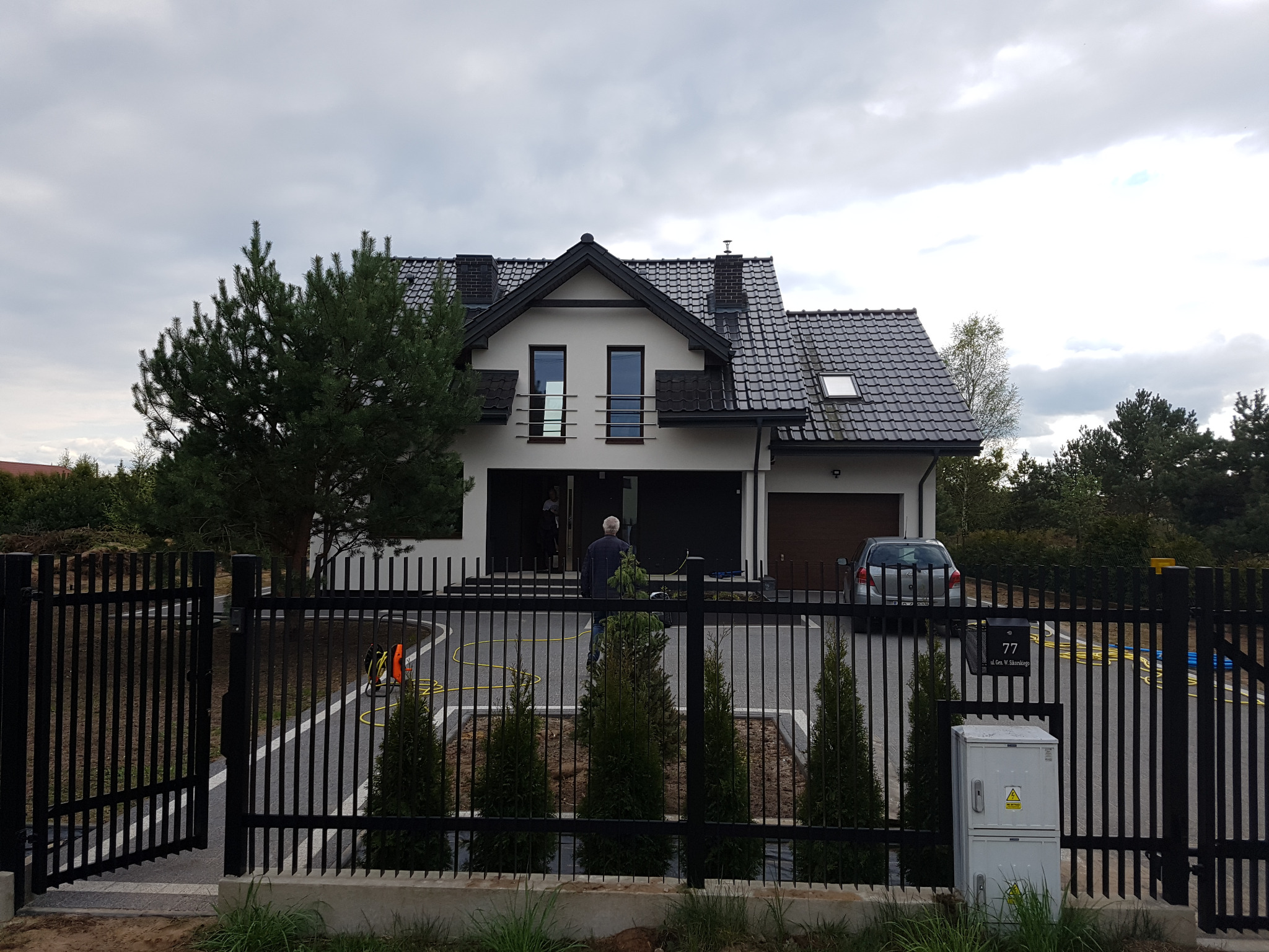 Nowoczesny dom jednorodzinny z czarnym dachem i białymi ścianami, widoczny przez czarne metalowe ogrodzenie z otwartą bramą. Na podjeździe stoi srebrny samochód, a w tle widać drzewa.