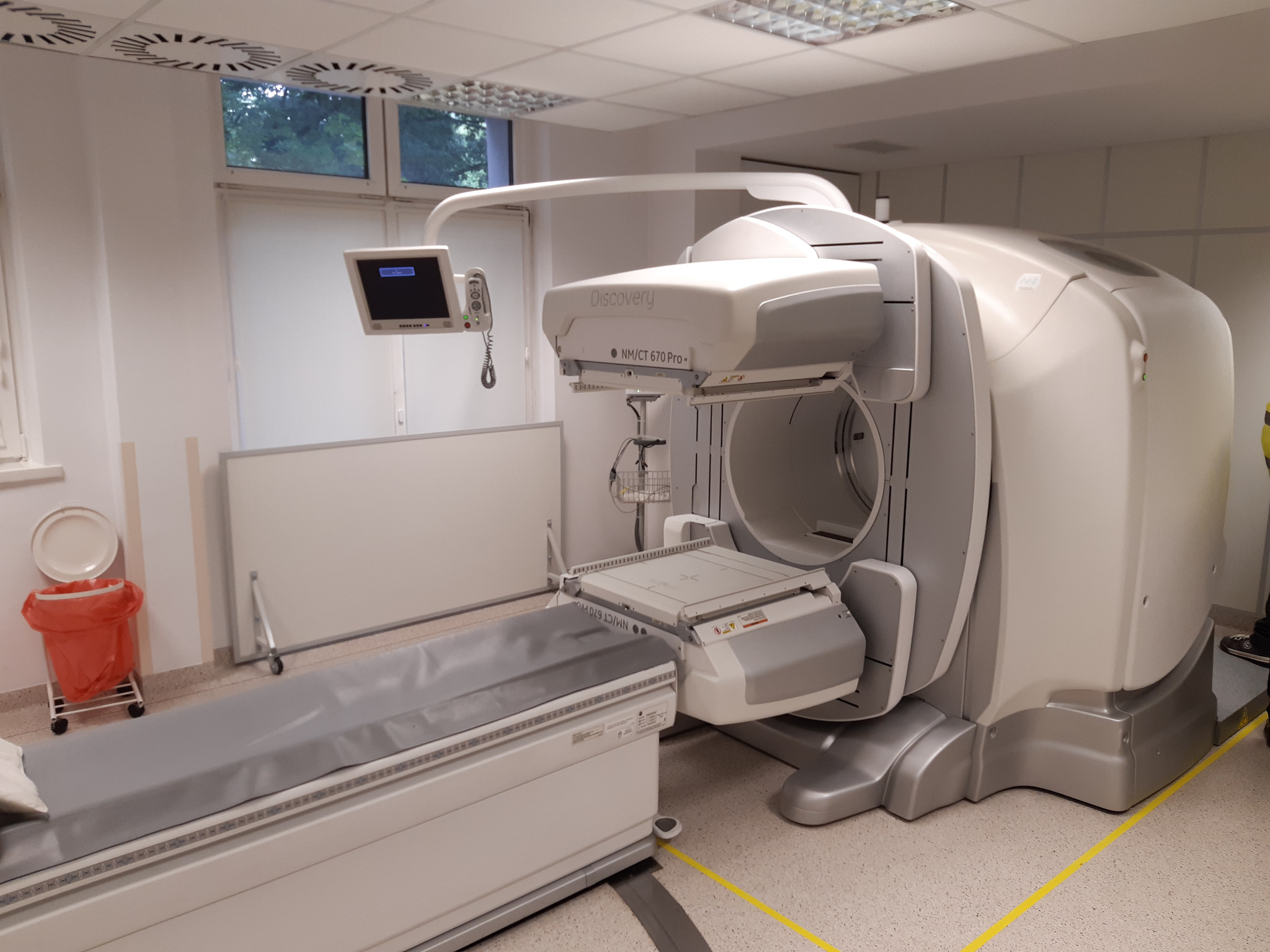 Aparat diagnostyczny Discovery NM/CT 670 Pro w jasnym pomieszczeniu, widoczne okno, kosz na śmieci i podłoga z terazzo.