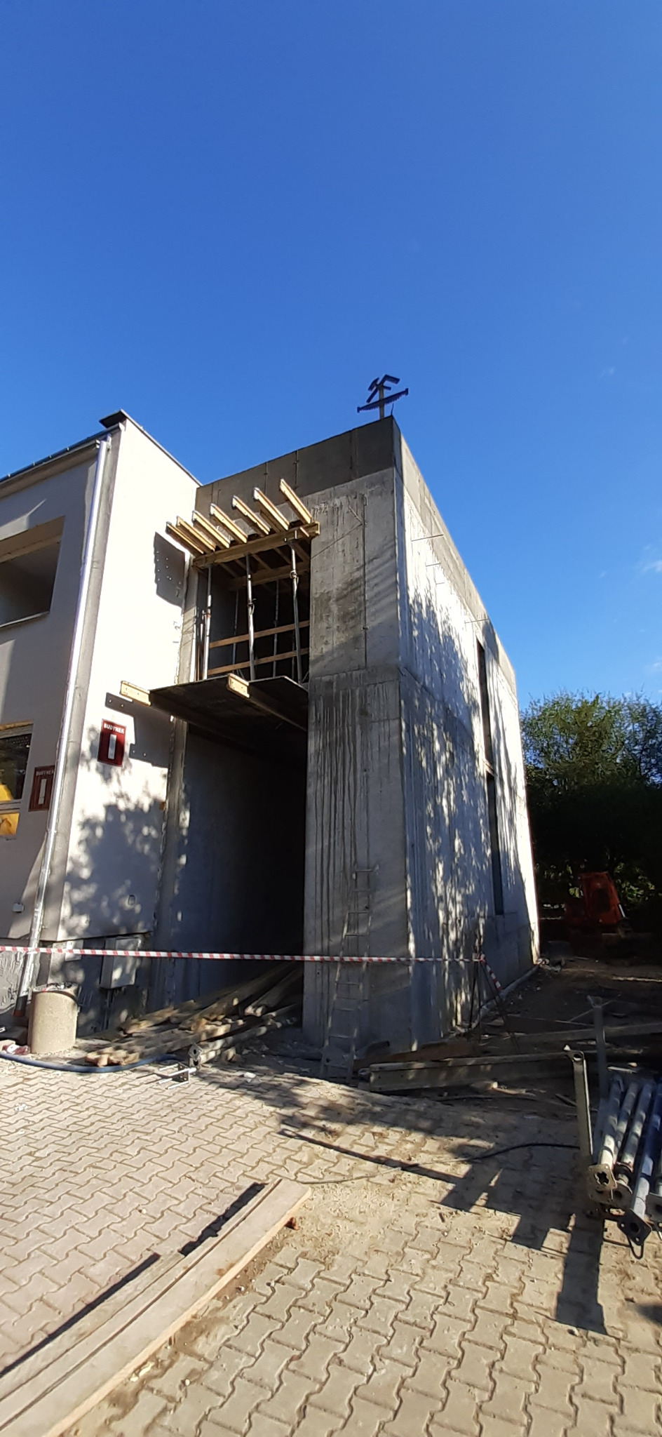 Surowy stan rozbudowy domu murowanego: betonowa konstrukcja połączona ze starszą częścią budynku, widoczne drewniane szalunki i zbrojenia, na dachu metalowa ozdoba, teren budowy zabezpieczony taśmą.