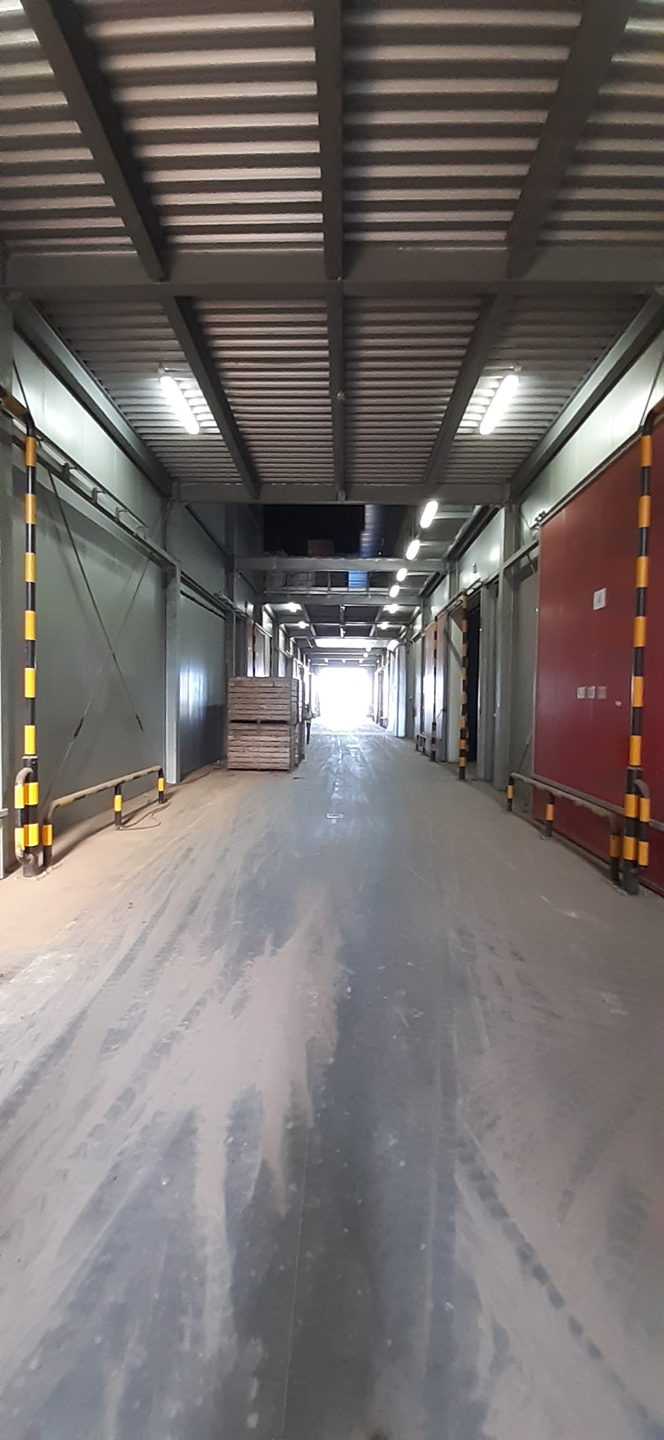 Wnętrze hali produkcyjnej z metalową konstrukcją dachu, oświetleniem jarzeniowym, betonową posadzką pokrytą pyłem i paletami składowanymi w głębi, widok w perspektywie z silnym światłem na końcu...