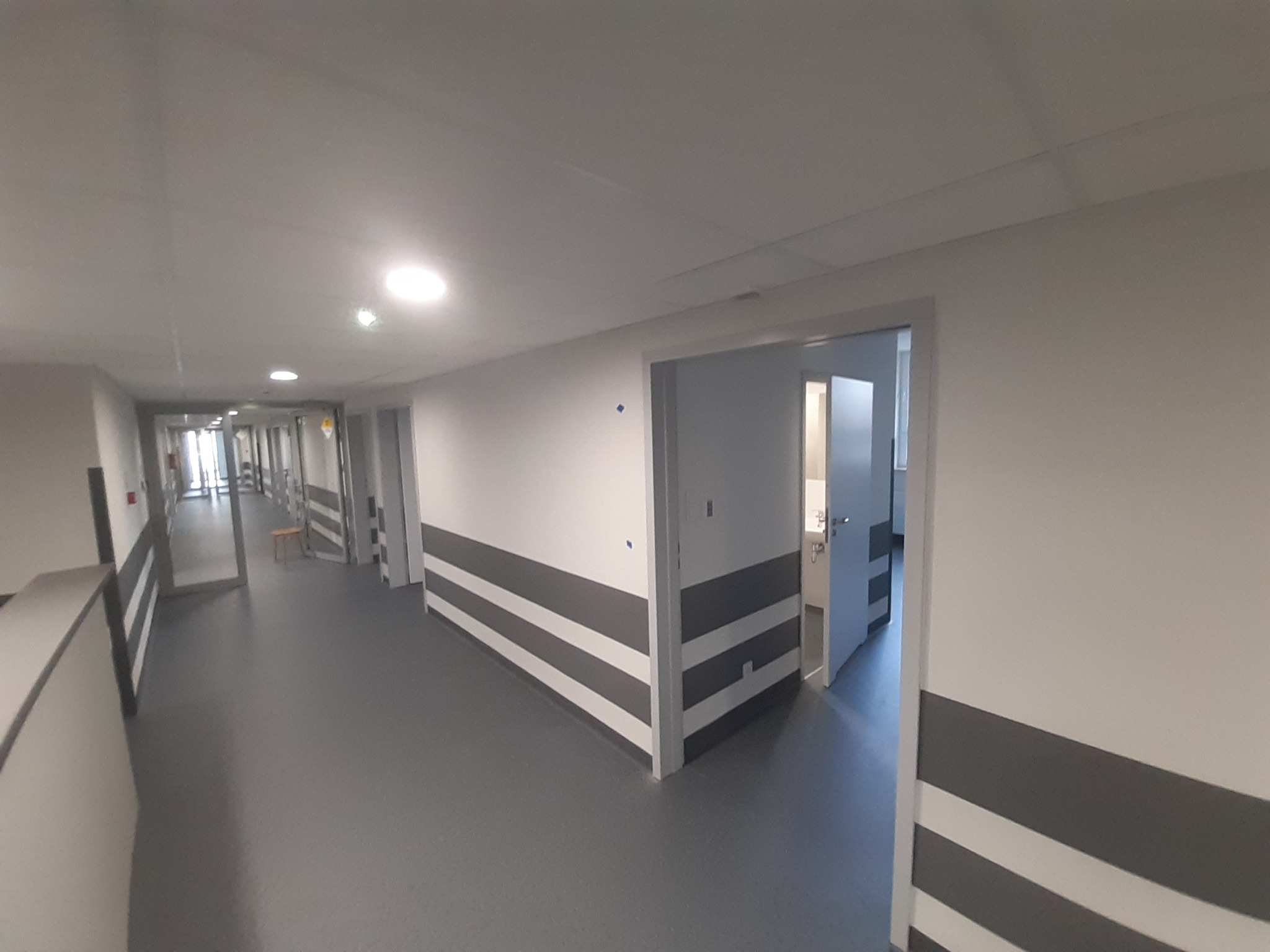 Długi korytarz wewnątrz budynku z szarymi pasami na ścianach i szarą podłogą, oświetlony lampami sufitowymi i z otwartymi drzwiami do pomieszczeń po bokach.