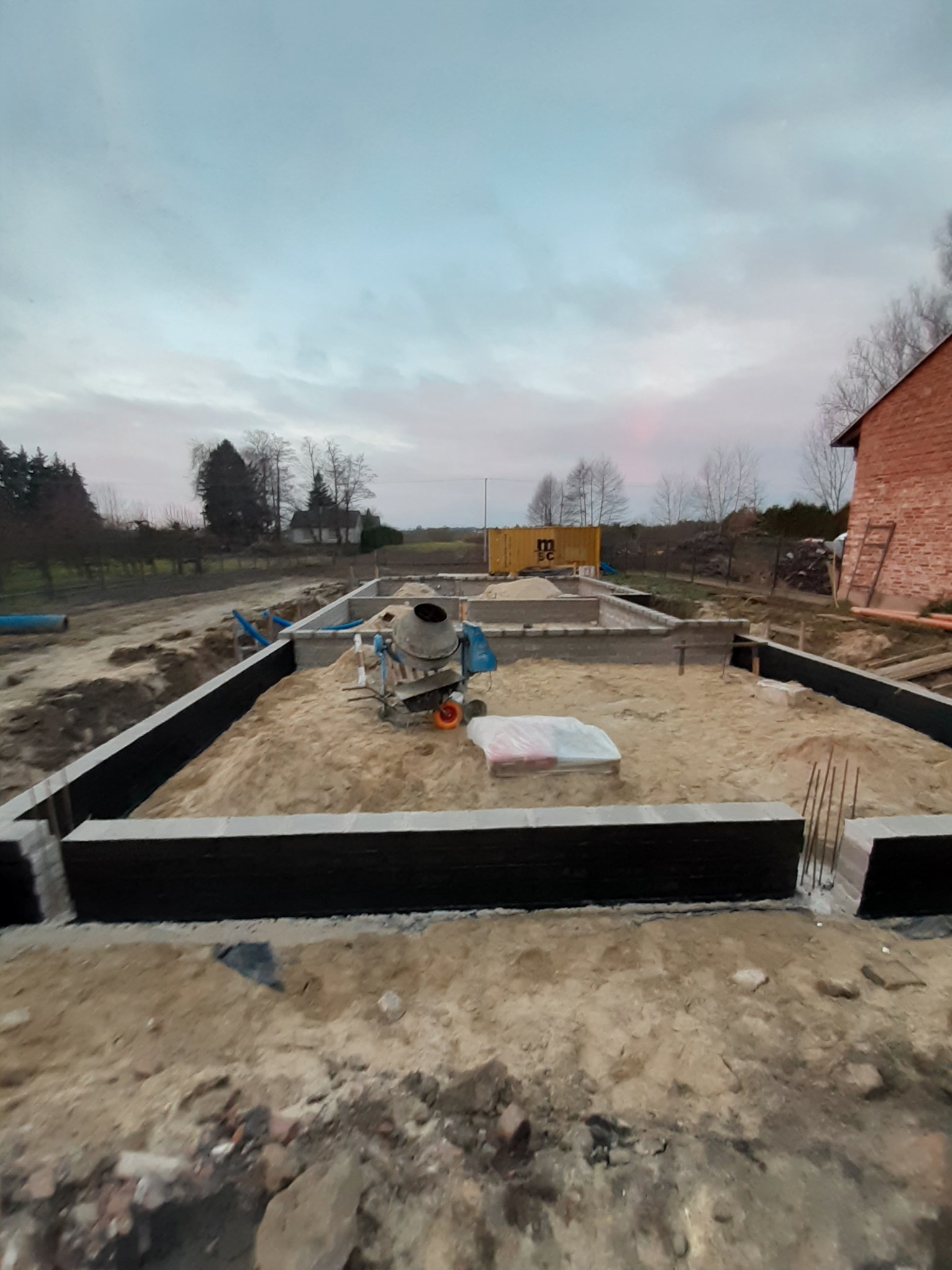 Przygotowany teren pod budowę domu z betonowymi fundamentami, widoczna betoniarka i materiały budowlane, w tle kontener i ceglany budynek gospodarczy, pochmurne niebo.