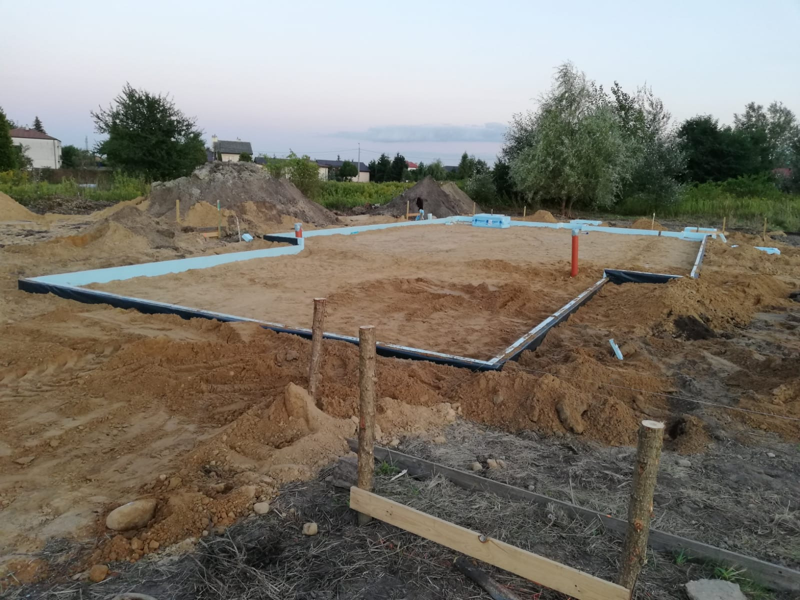 Przygotowany teren pod wylanie fundamentów domu, widoczne szalunki i izolacja termiczna, w tle hałdy piasku i drzewa.