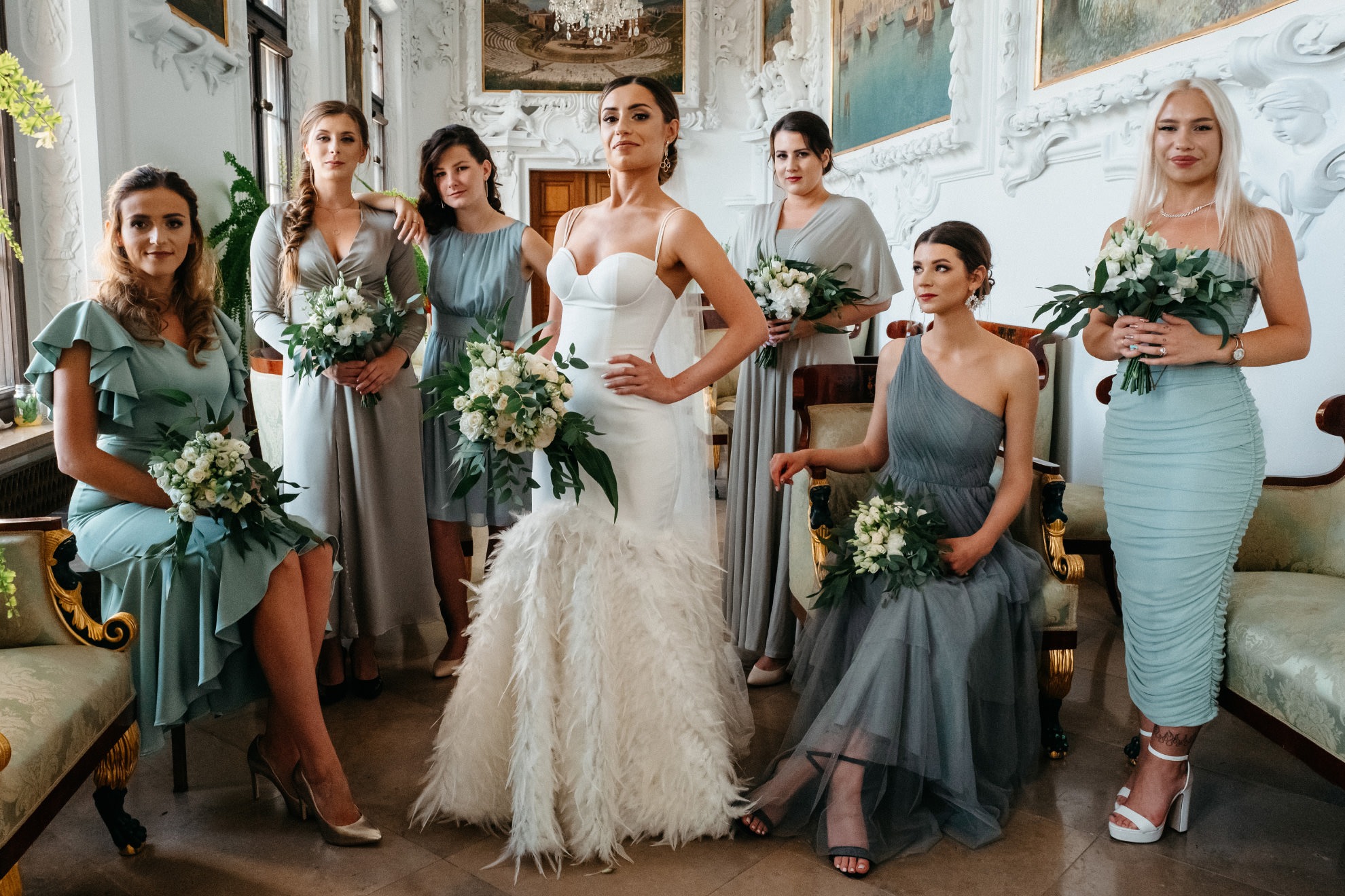 Panna młoda w białej sukni z piórami i druhny w pastelowych sukienkach pozują z bukietami na tle bogato zdobionego wnętrza pałacowego.