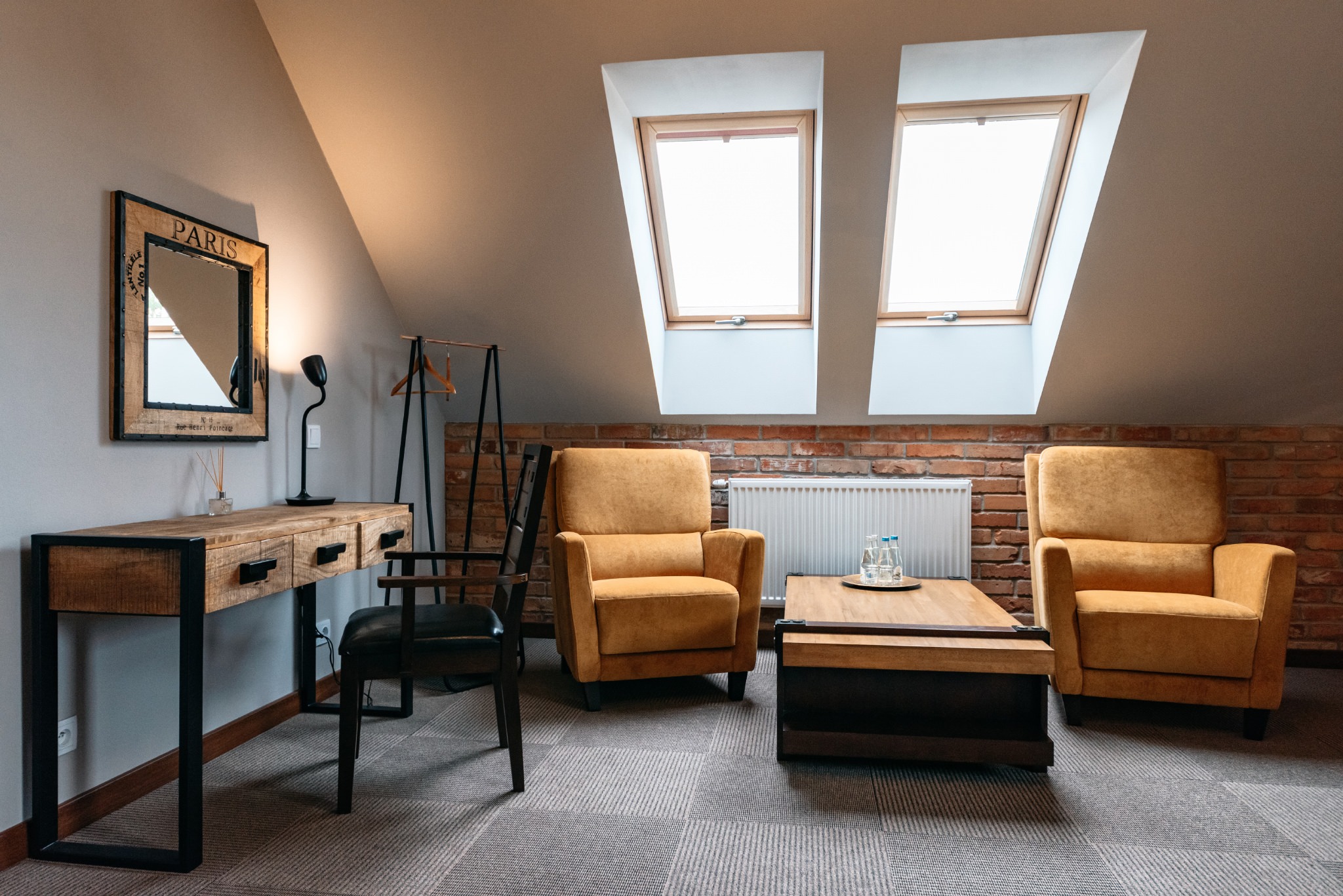 Pokój na poddaszu z dwoma oknami dachowymi, ceglaną ścianą, parą żółtych foteli, drewnianym stolikiem kawowym z wodą w butelkach, biurkiem z lustrem w stylu vintage i czarnym krzesłem.