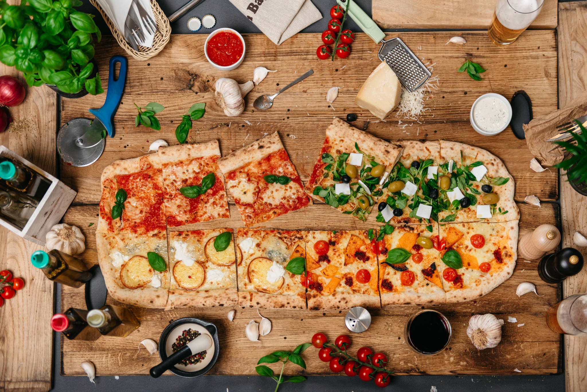Długa pizza z różnymi smakami na drewnianym stole, otoczona bazylią, oliwą, czosnkiem, pomidorami i przyprawami.