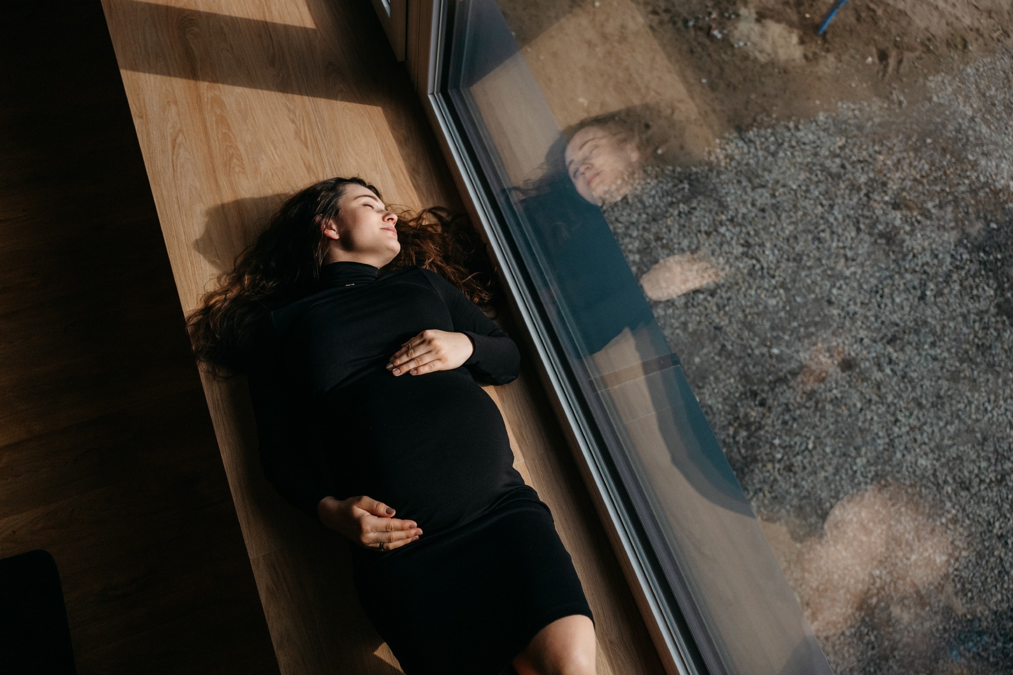 Kobieta w czarnej sukience ciążowej leży na drewnianej podłodze w promieniach słońca, z odbiciem w oknie ukazującym żwirowe podłoże.