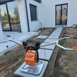 NIKO-BRUK - Układanie ścieżki z jasnoszarej kostki brukowej przy użyciu pomarańczowego urządzenia do przenoszenia. Widoczny fragment domu z dużymi oknami i przygotowany teren.