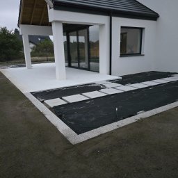 NIKO-BRUK - Nowoczesny taras w trakcie budowy: jasne płyty tarasowe, obrzeża z kostki granitowej, czarna geowłóknina. Widok na dom z dużymi oknami.