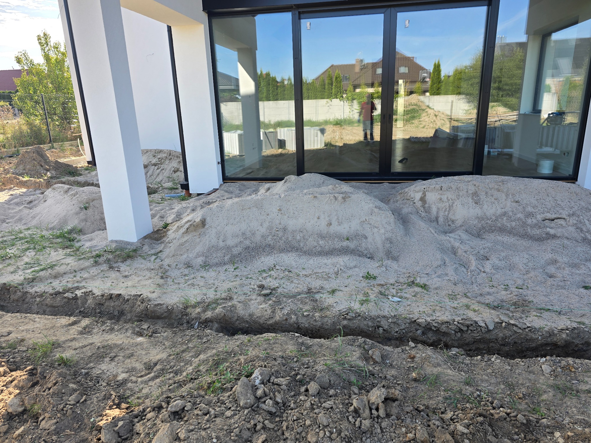 Przygotowanie terenu pod kostkę brukową: wykopany rów, usypany piasek przy nowoczesnym domu z dużymi oknami. W oknach odbija się osoba i otoczenie.