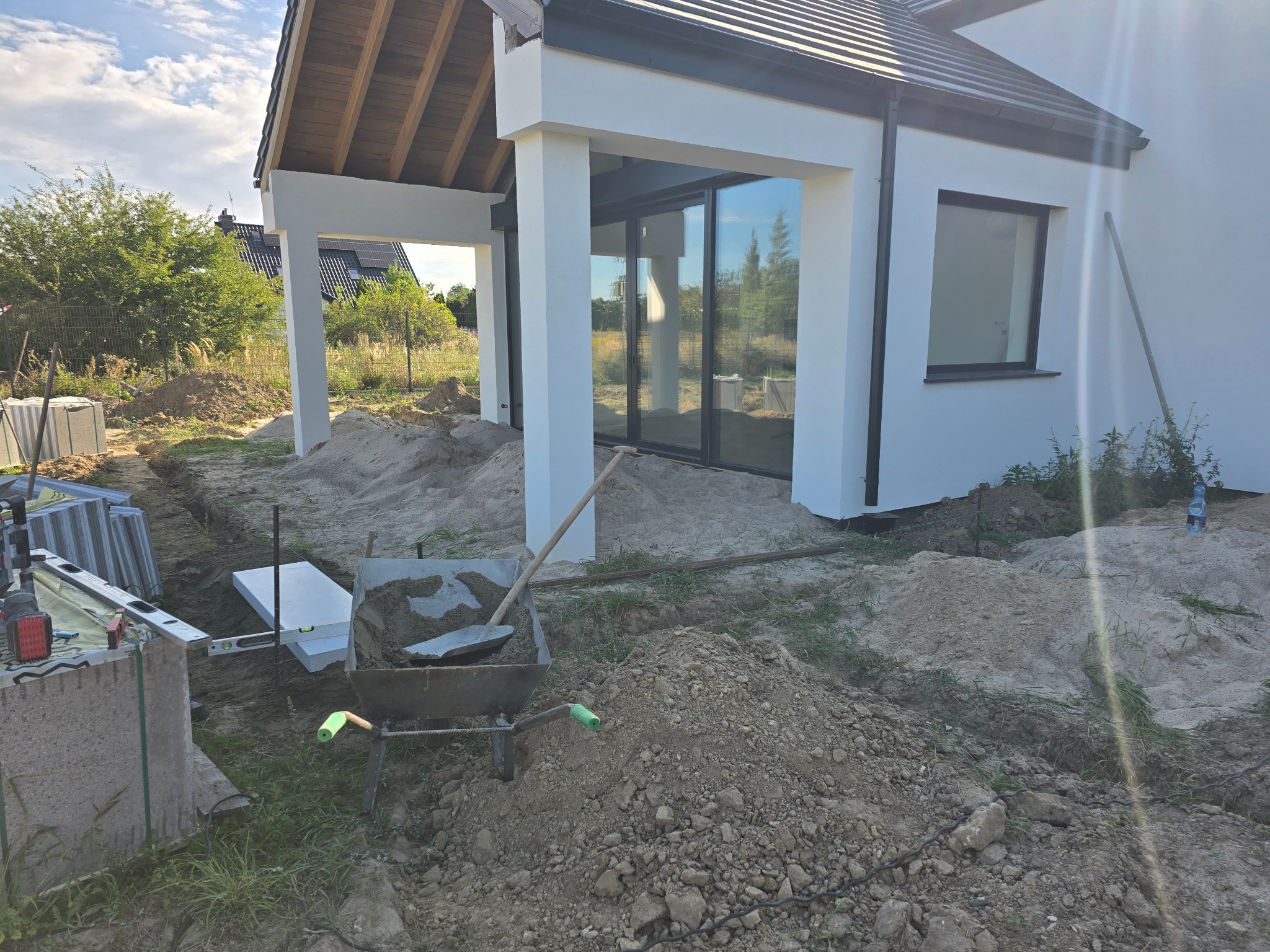 Przygotowanie terenu pod kostkę brukową przy nowoczesnym domu z białymi kolumnami. Widoczna taczka z piaskiem, poziomica i ułożone obrzeża. Etap prac ziemnych.