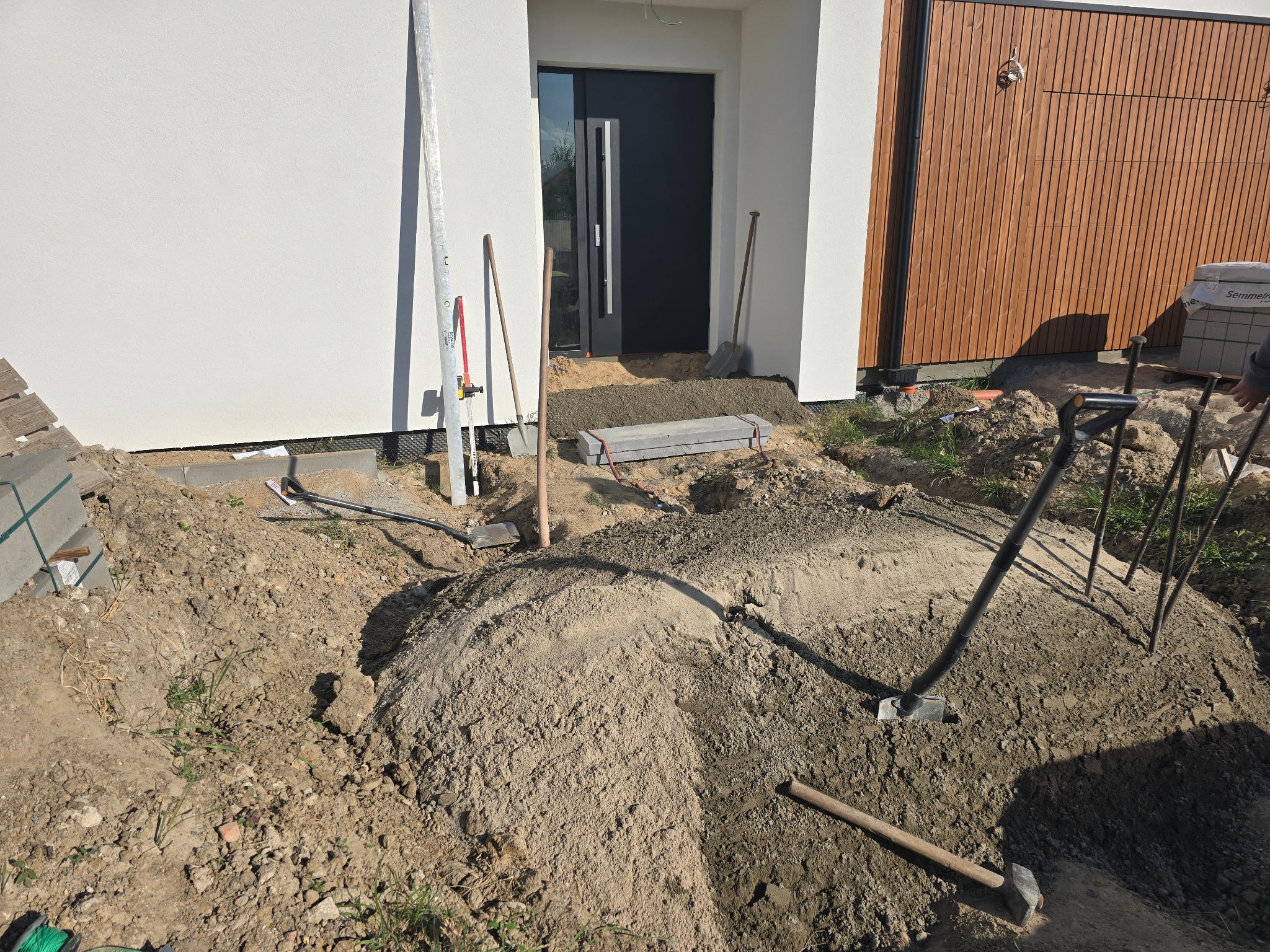 Przygotowanie podłoża pod kostkę brukową przed nowoczesnym domem z drewnianą elewacją. Widoczne narzędzia: łopaty, poziomica, młotek i kostka brukowa.