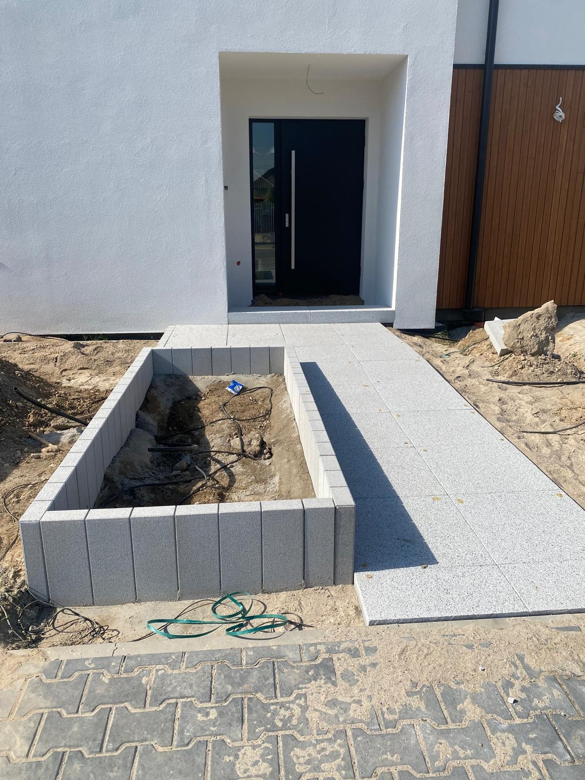 Jasnoszara ścieżka z płyt prowadząca do drzwi wejściowych, obok rabata z obrzeżem z kostki. Widoczne kable w ziemi. Etap prac przed wykończeniem.