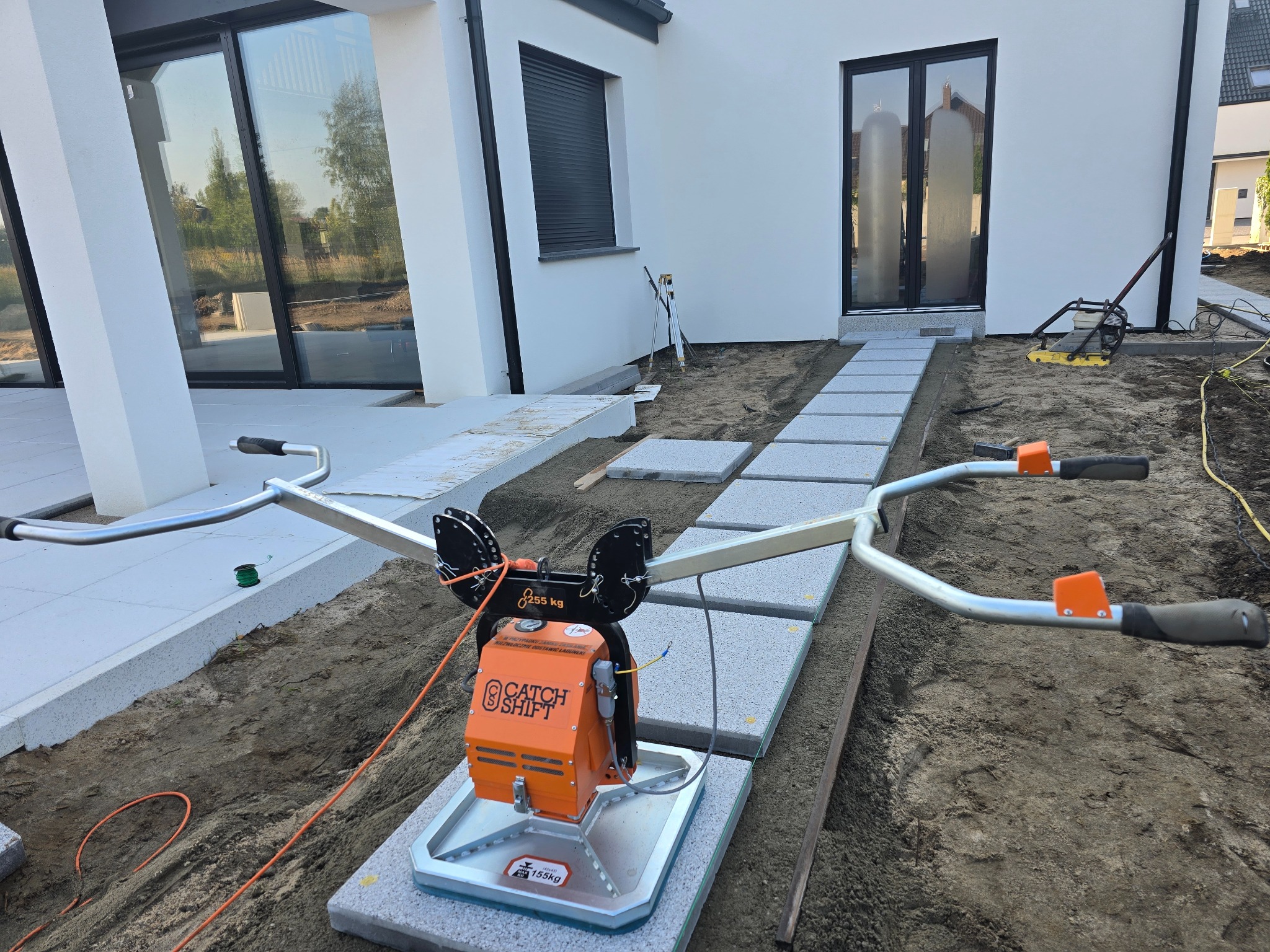 Układanie ścieżki z jasnoszarej kostki brukowej przy użyciu pomarańczowego urządzenia do przenoszenia. Widoczny fragment domu z dużymi oknami i przygotowany teren.