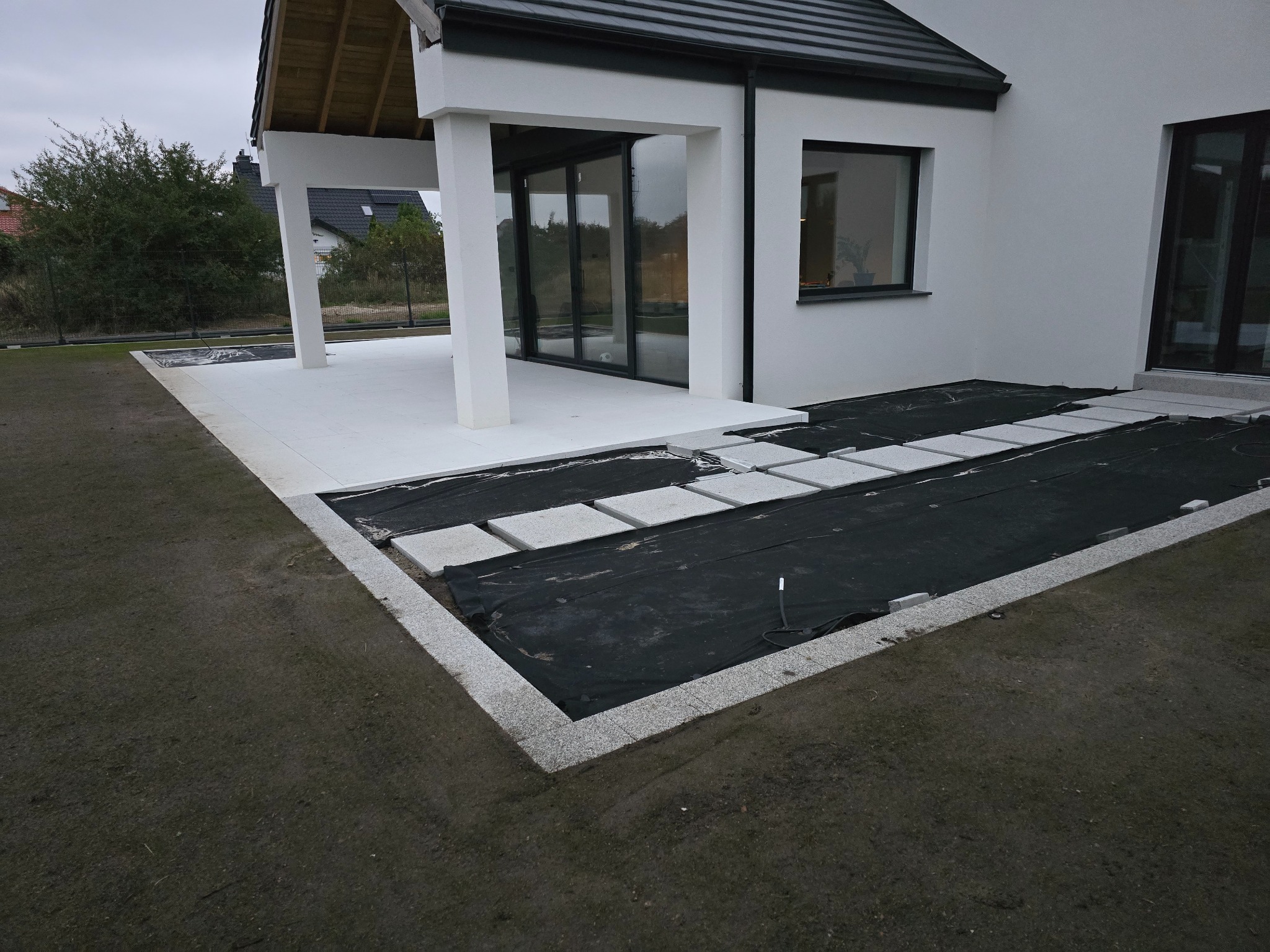 Nowoczesny taras w trakcie budowy: jasne płyty tarasowe, obrzeża z kostki granitowej, czarna geowłóknina. Widok na dom z dużymi oknami.