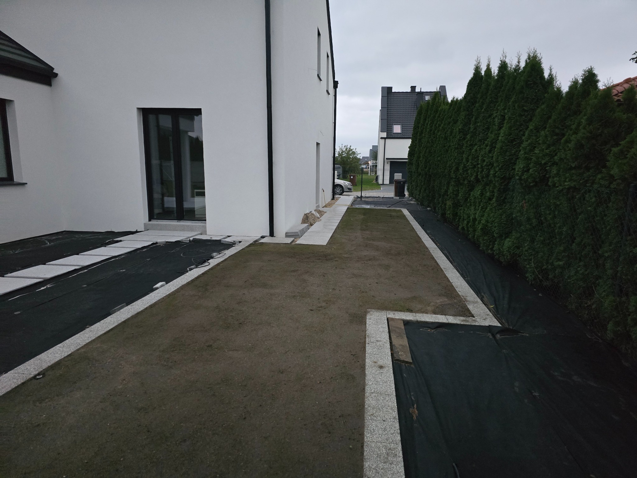 Przygotowanie terenu pod ścieżkę z płyt i obrzeży w Ostrorogu. Widoczna czarna agrowłóknina i jasne płyty. Minimalistyczny styl domu i wysokie tuje w tle.