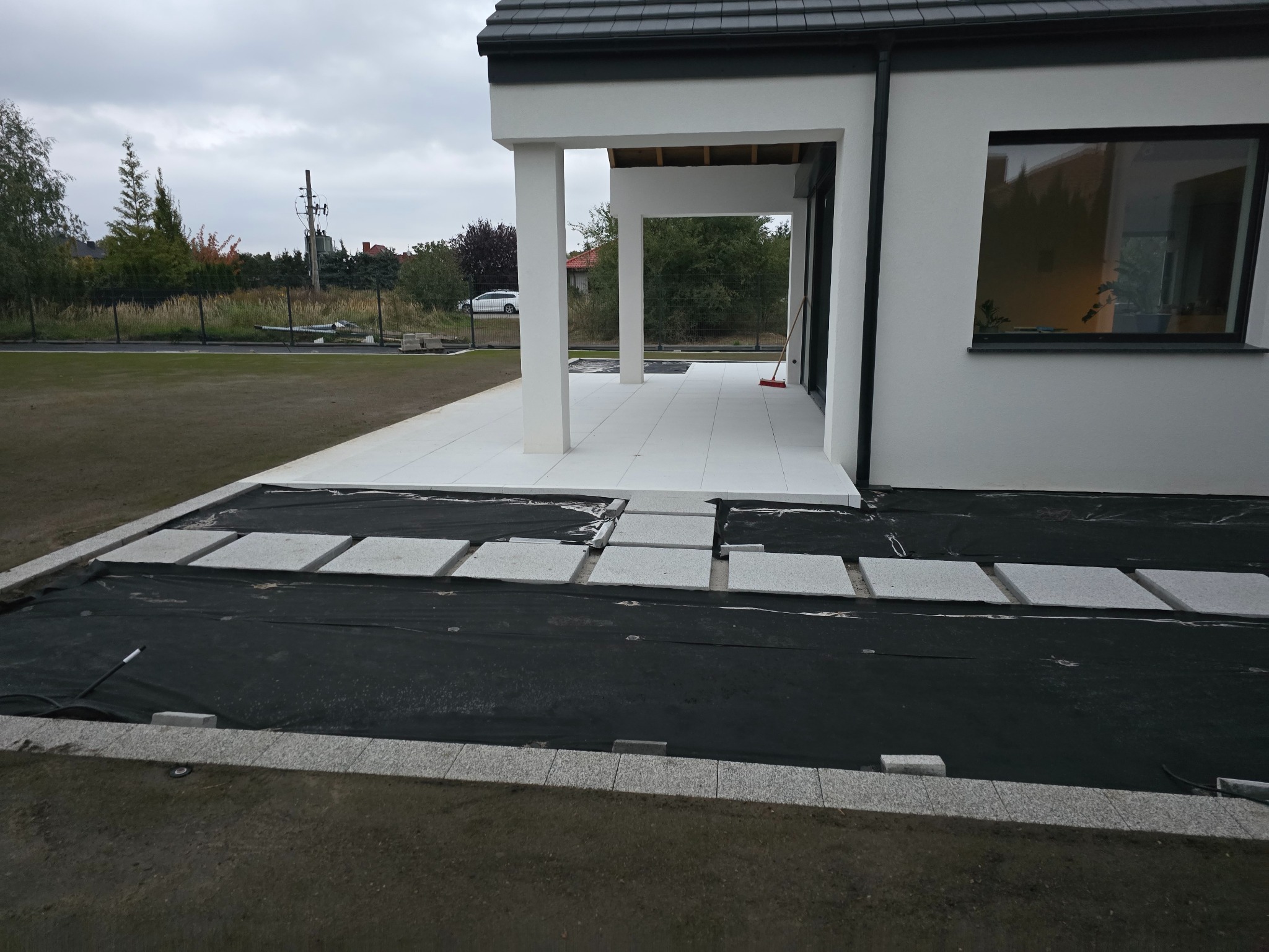 Nowoczesny taras z jasnych płyt i kostki granitowej w trakcie realizacji. Widoczna czarna geowłóknina i obrzeża. Dom z białymi kolumnami w tle.