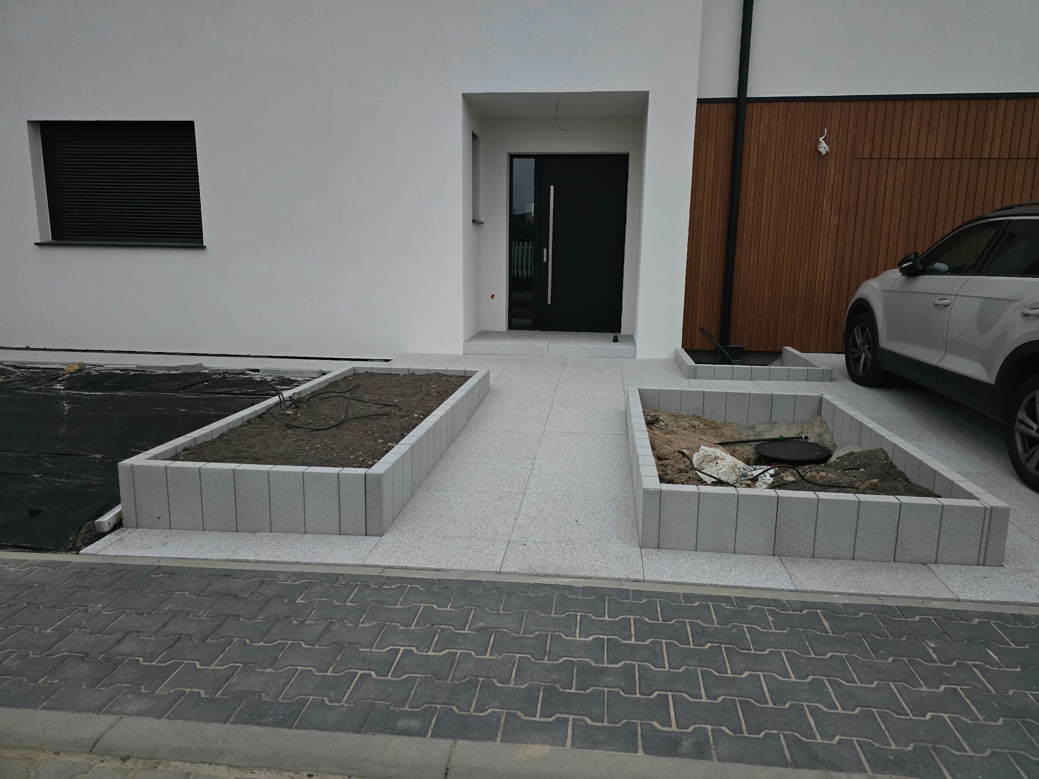 Nowoczesny podjazd z jasnoszarej kostki granitowej i obrzeżami, prowadzący do domu z czarnymi drzwiami. Widoczne rabaty z kostki i fragment kostki brukowej.