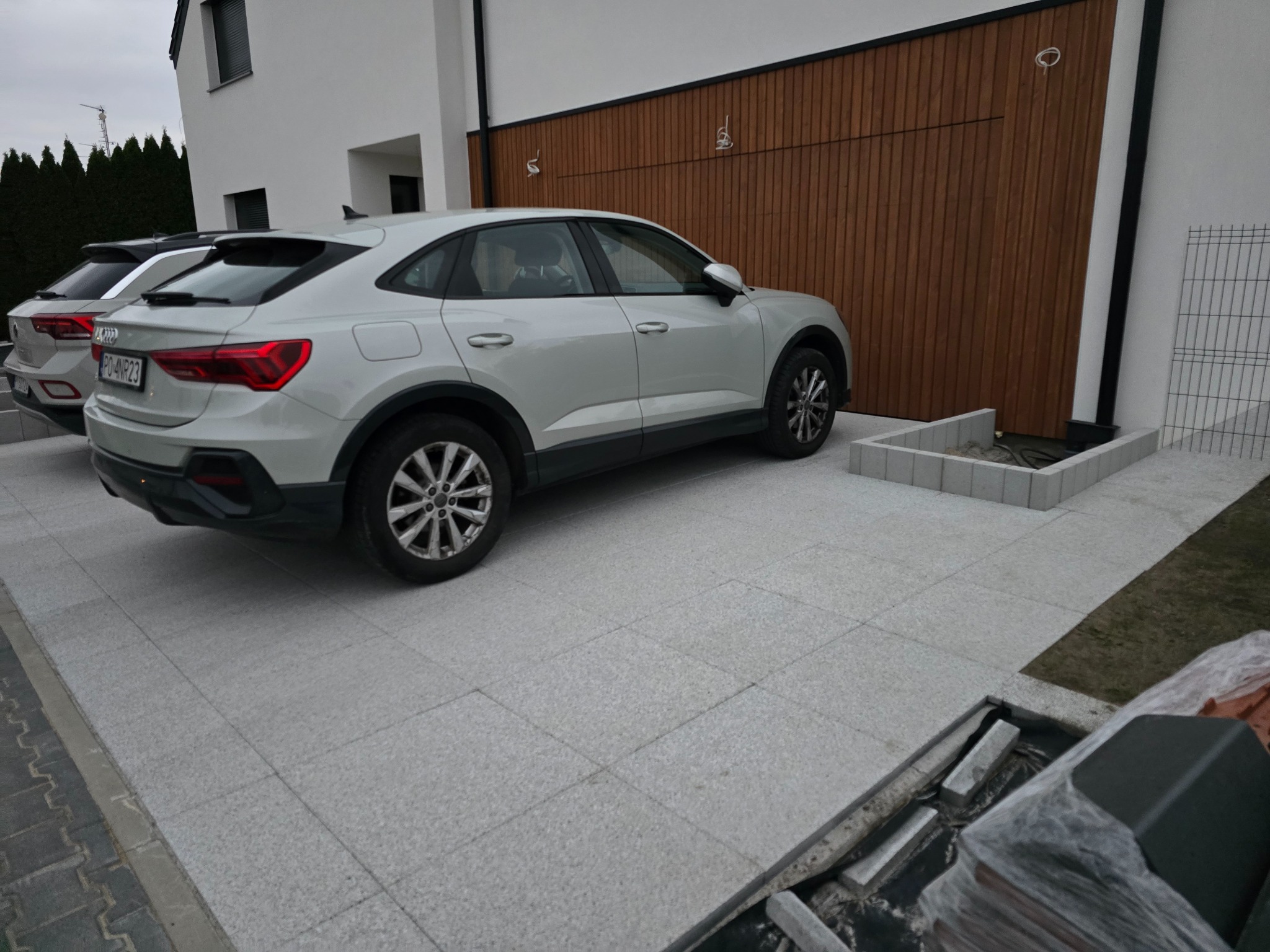 Jasnoszara kostka brukowa ułożona na podjeździe przed nowoczesnym domem z drewnianą bramą garażową. Zaparkowane dwa samochody Audi.