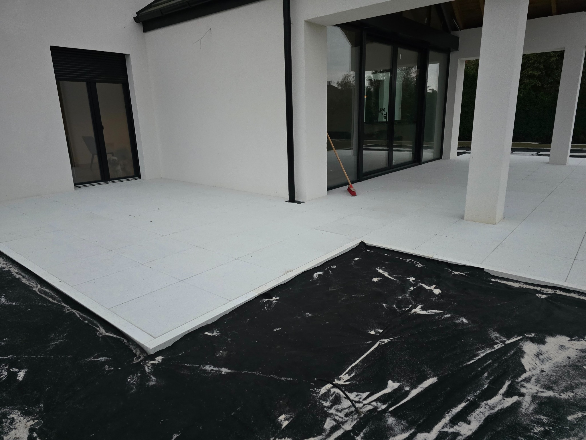 Biała, nowoczesna kostka na tarasie domu w trakcie układania. Fragment z czarną folią i piaskiem. Widoczny budynek z oknami i kolumnami. Czysta, minimalistyczna estetyka.