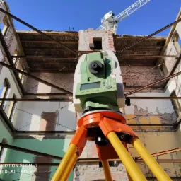 Tachimetr na statywie ustawiony wewnątrz ruin budynku z widocznym żurawiem budowlanym na tle błękitnego nieba.