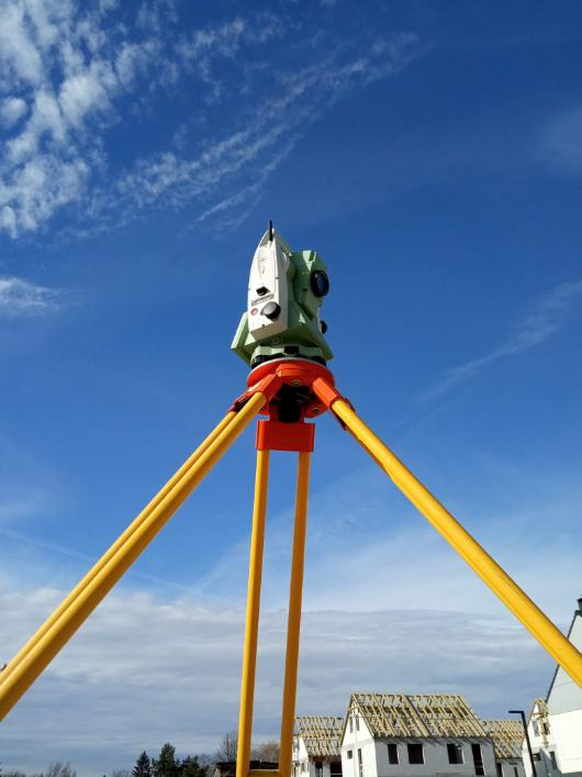 Teodolit na statywie z żółtymi nogami na tle błękitnego nieba z chmurami, w tle budynki w trakcie budowy z widoczną konstrukcją dachu.