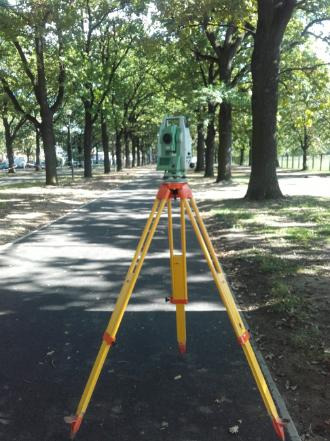 Teodolit na żółtym statywie ustawiony na ścieżce w parku, w tle szpaler drzew.