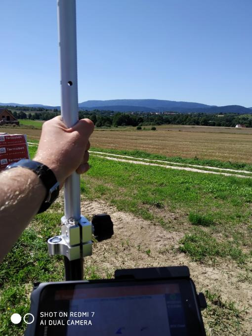 Praca w terenie: osoba trzyma tyczkę geodezyjną z odbiornikiem GPS, w tle zielone pola i zalesione wzgórza w oddali.