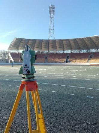 Praca pomiarowa teodolitem na stadionie w Legnicy, w tle widoczna konstrukcja oświetleniowa i zadaszenie trybun.