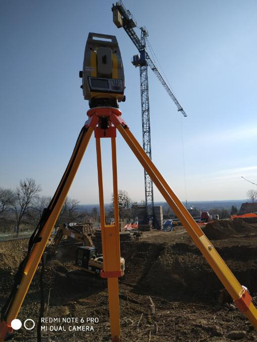 Żółty teodolit na statywie ustawiony na placu budowy z koparką i żurawiem w tle, błękitne niebo.