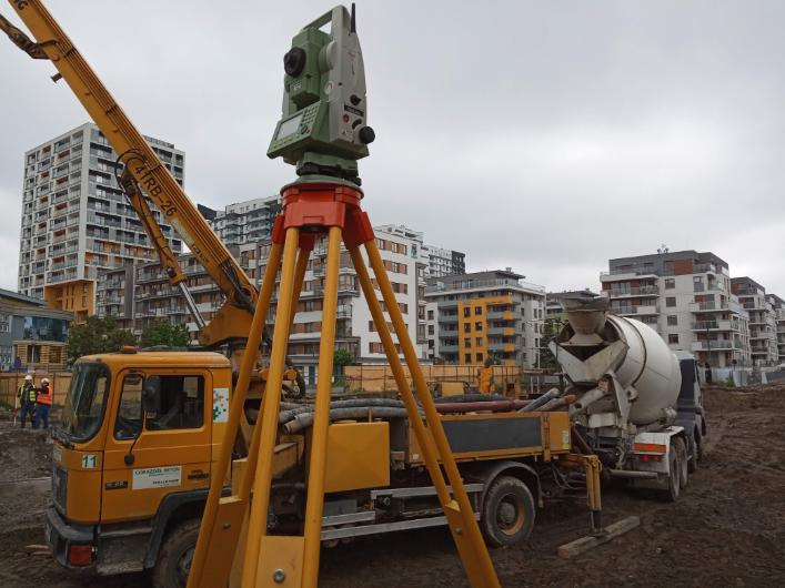 Żółty teodolit na trójnogu ustawiony na tle budowy z betoniarką i budynkami mieszkalnymi w tle, pochmurne niebo.