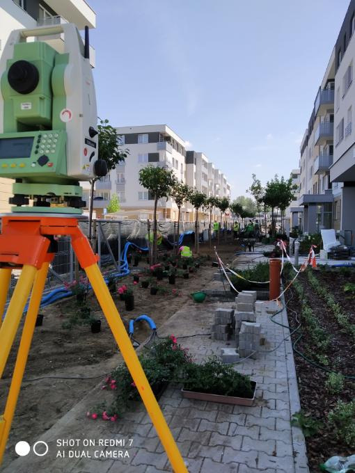 Pomiar geodezyjny terenu przed budynkami mieszkalnymi, z widocznym teodolitem na trójnogu, doniczkami z kwiatami i ułożonymi kostkami brukowymi.
