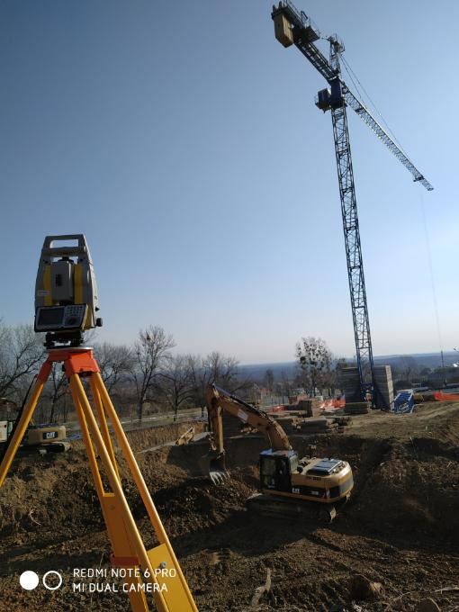 Żółty teodolit na trójnogu na pierwszym planie, w tle plac budowy z koparkami CAT i wysokim dźwigiem wieżowym na tle błękitnego nieba.