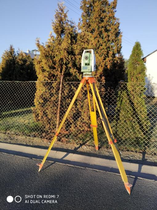 Zielony teodolit na żółtym statywie ustawiony na asfalcie, z widoczną metalową siatką ogrodzeniową i tujami w tle, w słoneczny dzień.